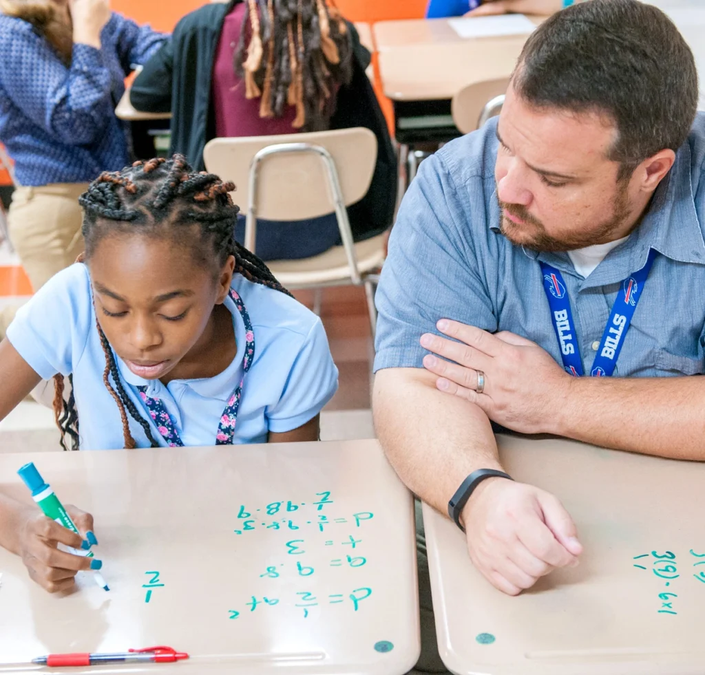 A teachers sits at a desk and helps a student who is practicing math problems using a dry-erase marker.