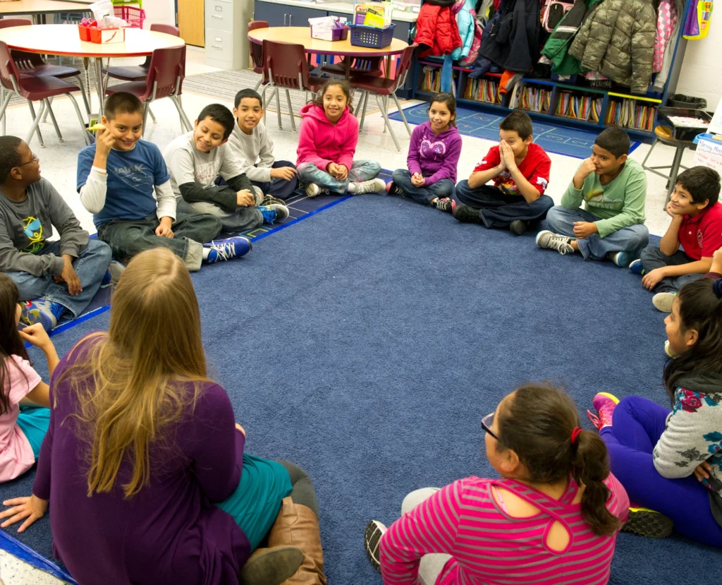 Students sit in a circle alongside their teacher as they enjoy a Morning Meeting sharing activity.