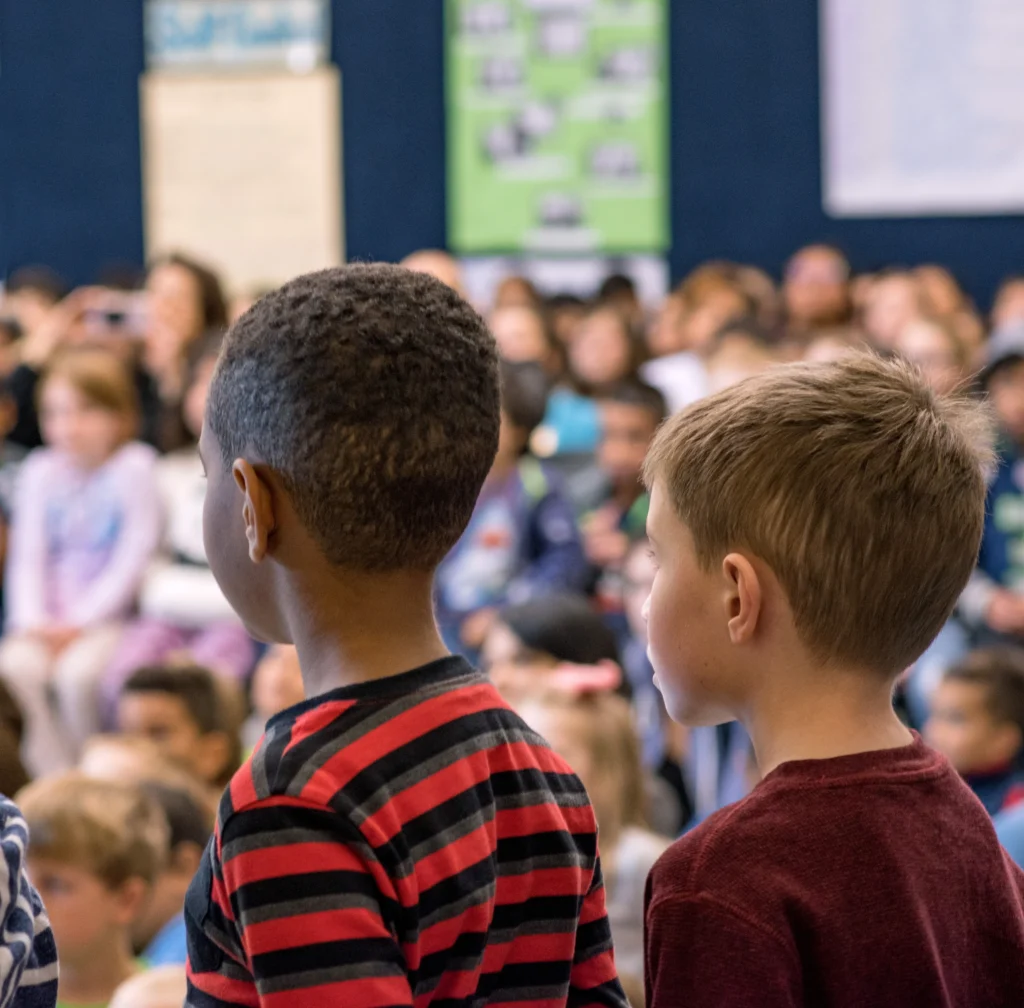 Two elementary school boys stand at the front of a school-wide assembly in the school gym.