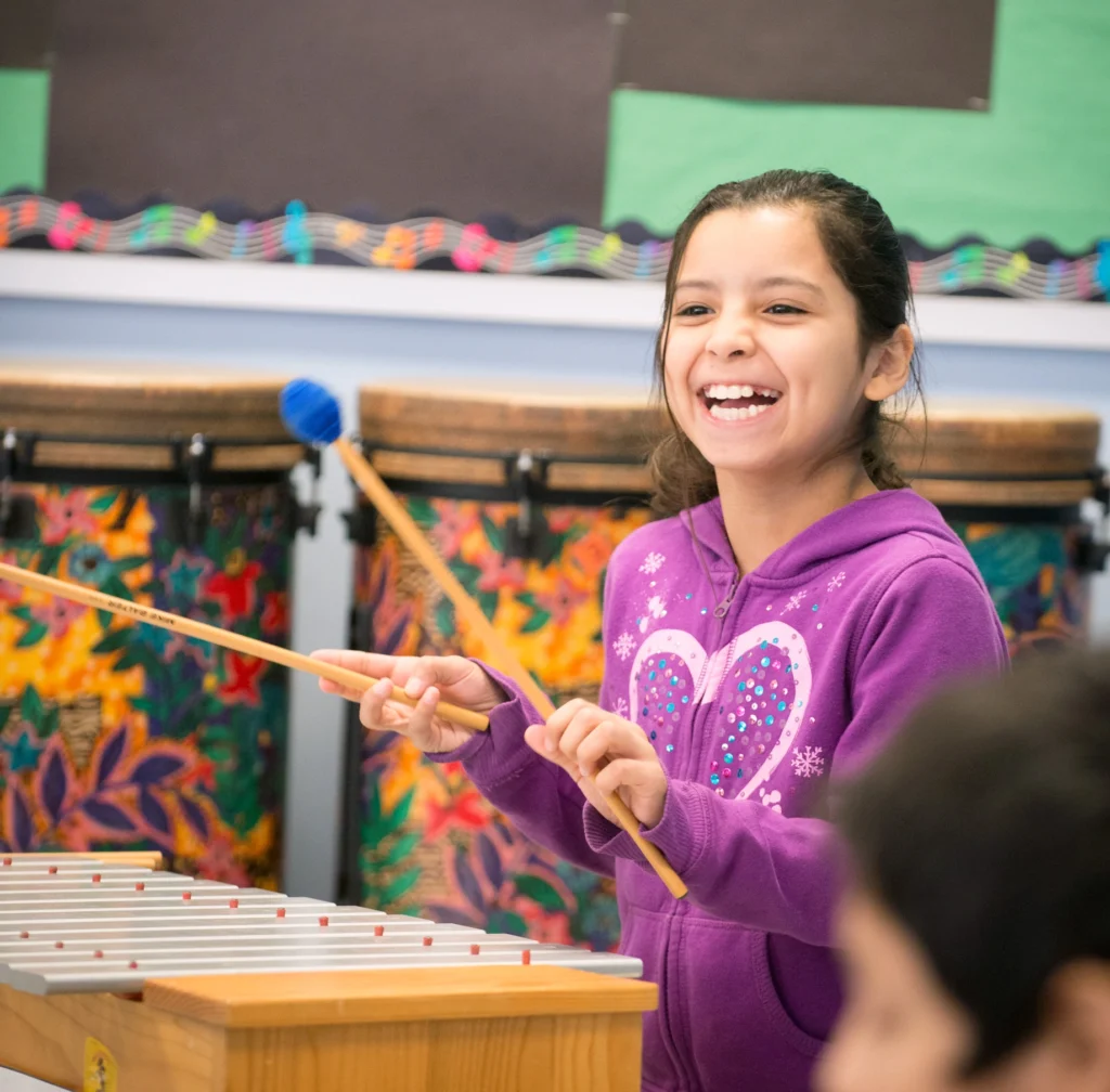 An young female elementary schools student is laughing while playing the xylophone in her special area teacher's music class.